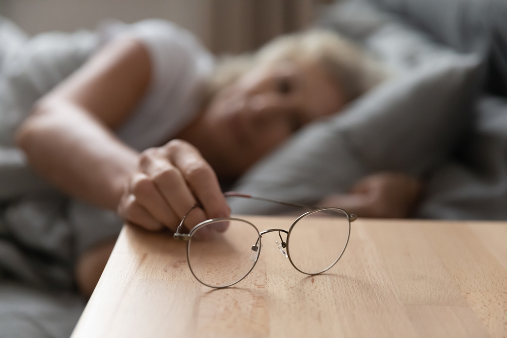 Close up focus on eyeglasses on wooden bedside table. Elderly senior retired woman suffering from blurred eyesight, taking optical eyewear after waking up in bedroom, bad vision ophthalmology concept.