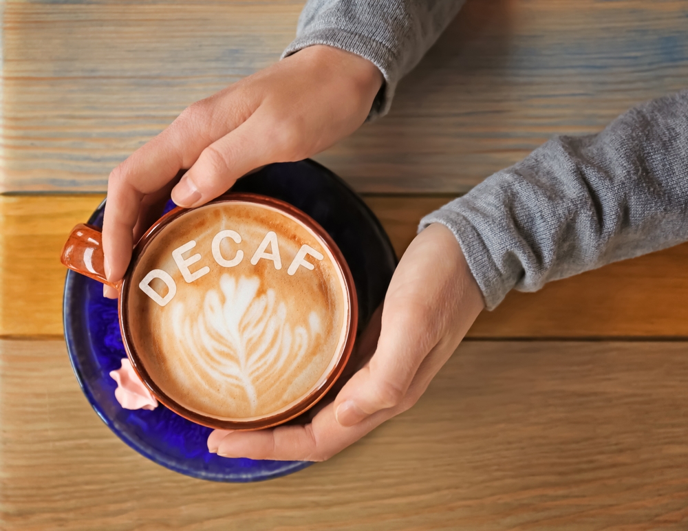 Woman holding cup of delicious decaf coffee with foam on wooden table, top view