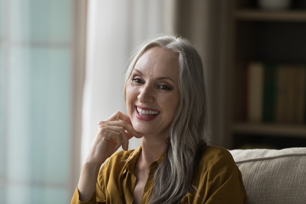 Happy cheerful pretty mature woman candid head shot portrait. Joyful beautiful long grey haired lady looking away with toothy smile, laughing, speaking, touching chin. Elderly age, beauty care