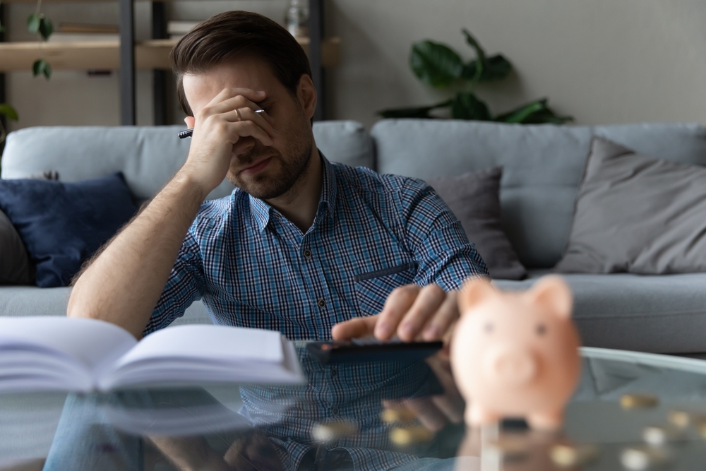 Man calculates expenses feels concerned due finances shortage, lack of money to pay for loan cover face with palm looks upset, blurred piggybank on table. Crisis, bankruptcy, financial problem