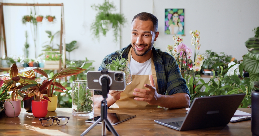 African American Blogger gardener uses phone while tending houseplants and using shovel on table. Concept of plant care and gardening for small busines. Spring planting. Social media.