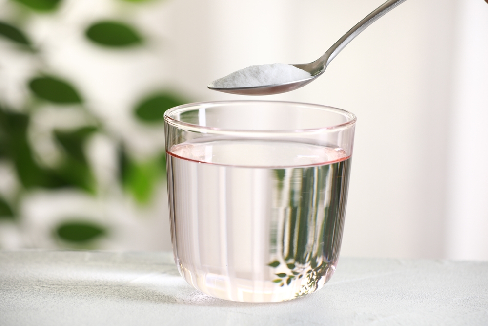 Spoon baking soda over a glass of water on a white table against a blurred background, close up