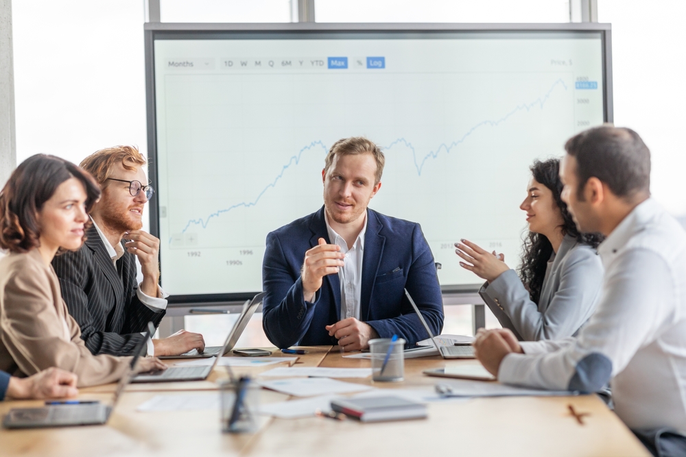 A group of business professionals are seated around a conference table, engaged in a discussion. They are gathered in a modern office with a large whiteboard behind them