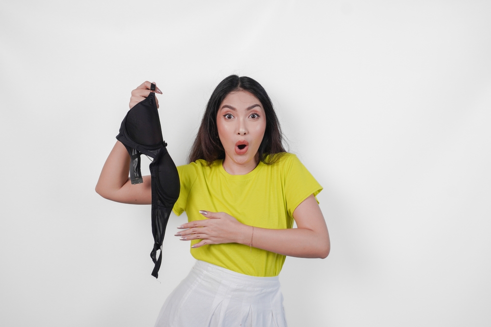 Surprised young Asian woman in yellow shirt is holding a black bra for no bra day celebration, isolated by white background.