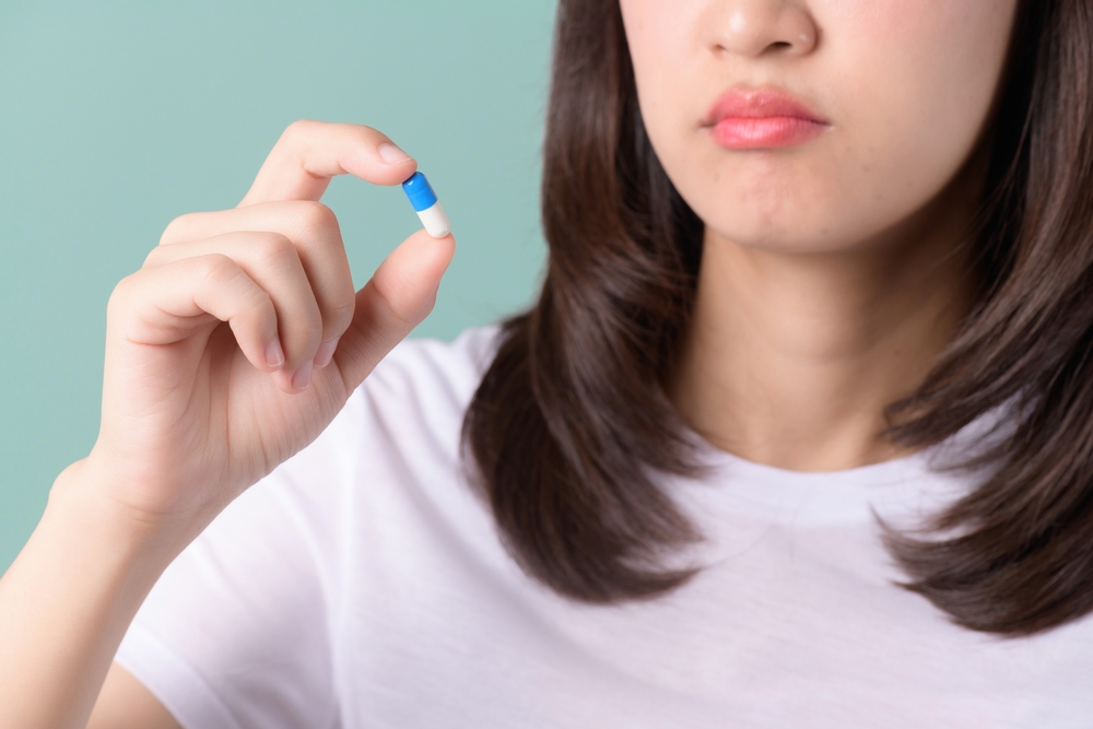 Forget taking medicine. A confused woman holds her head and looks at pills on a green field. An Asian woman forgets to take a medicine, misses a dose, and thinks about the next prescription with a pill in her hand.