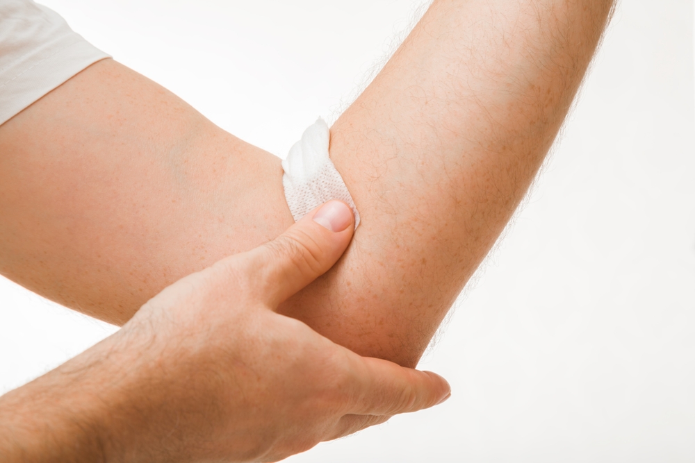 Young adult man gluing and putting bandage with cotton wool on arm vein after blood test on light gray background. Closeup. Front view.