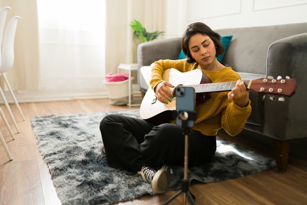 A creative musician practicing and recording music lessons at home, using a smartphone to record his acoustic guitar performance