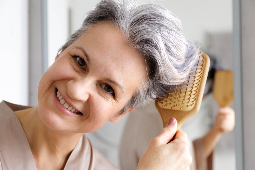 A smiling elderly beautiful woman in her retirement age in silk pajamas combs her gray hair.