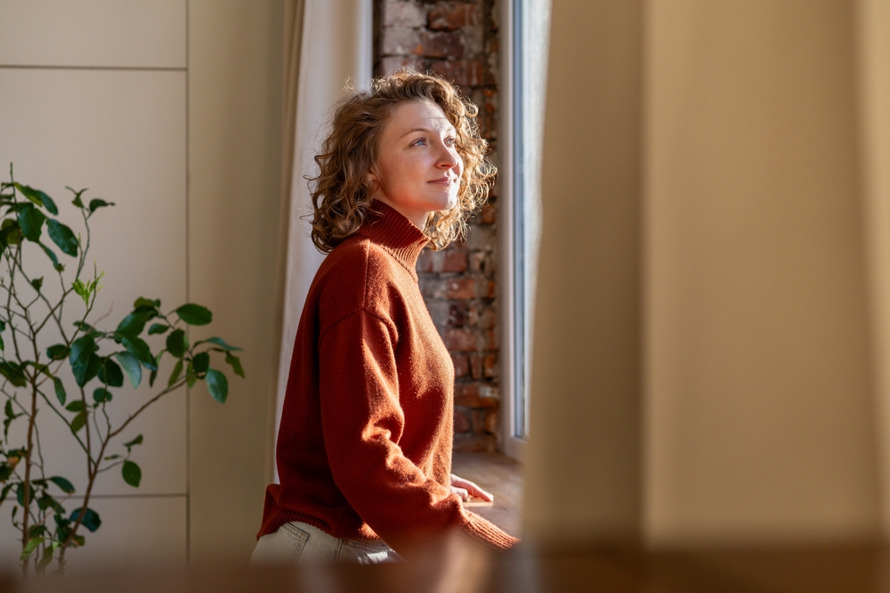 Positive thoughtful woman enjoys tranquil break at home, leaning by window feeling gentle sunlight on skin. Carefree young female with peaceful heart, joyful mind relishes beauty of day taking pause.