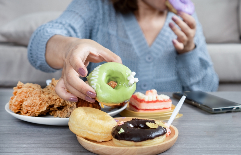 woman’s hand reaches for a donut on table that full of junk food like fried chicken, cake, sweet drinks, and desserts, showing signs of sugar addiction, eating too much, and eating unhealthy food 