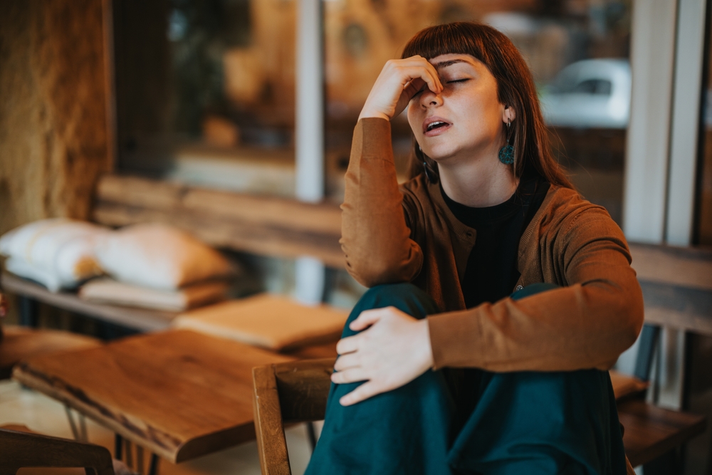 A sad woman sits at a coffee bar with a stressed expression. The cozy atmosphere contrasts with her emotions, highlighting feelings of depression and fatigue in a public space.