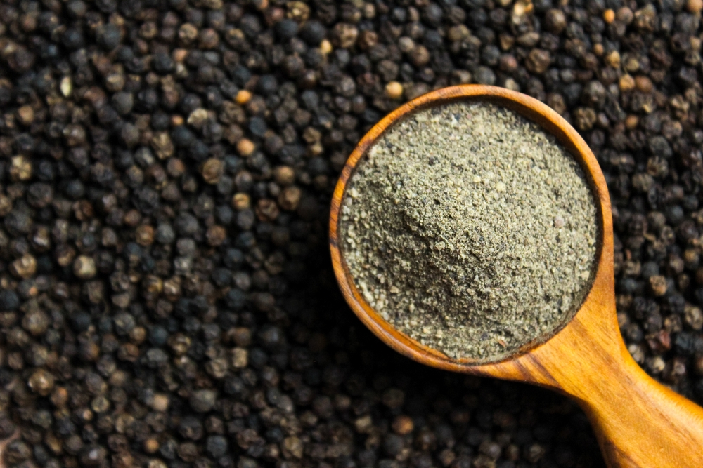 Black pepper powder in wooden cup spoon with black peppercorns background. Food ingredients. Aromatic spice. Top view