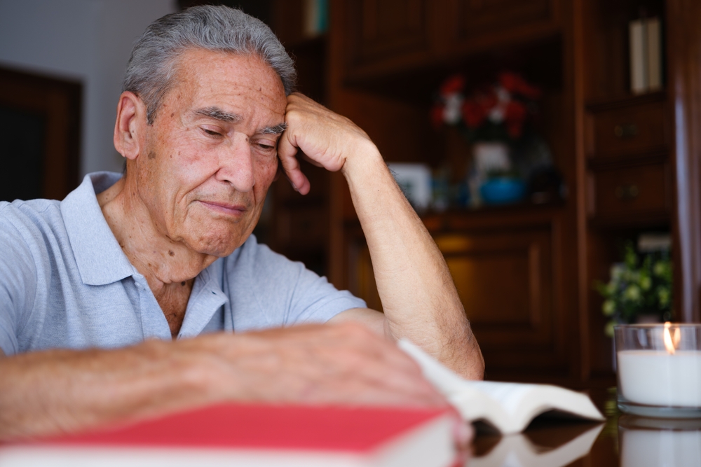 Elderly man reading at home with a lit candle, in a horizontal side view with a nostalgic expression, resting his head at a wooden table.