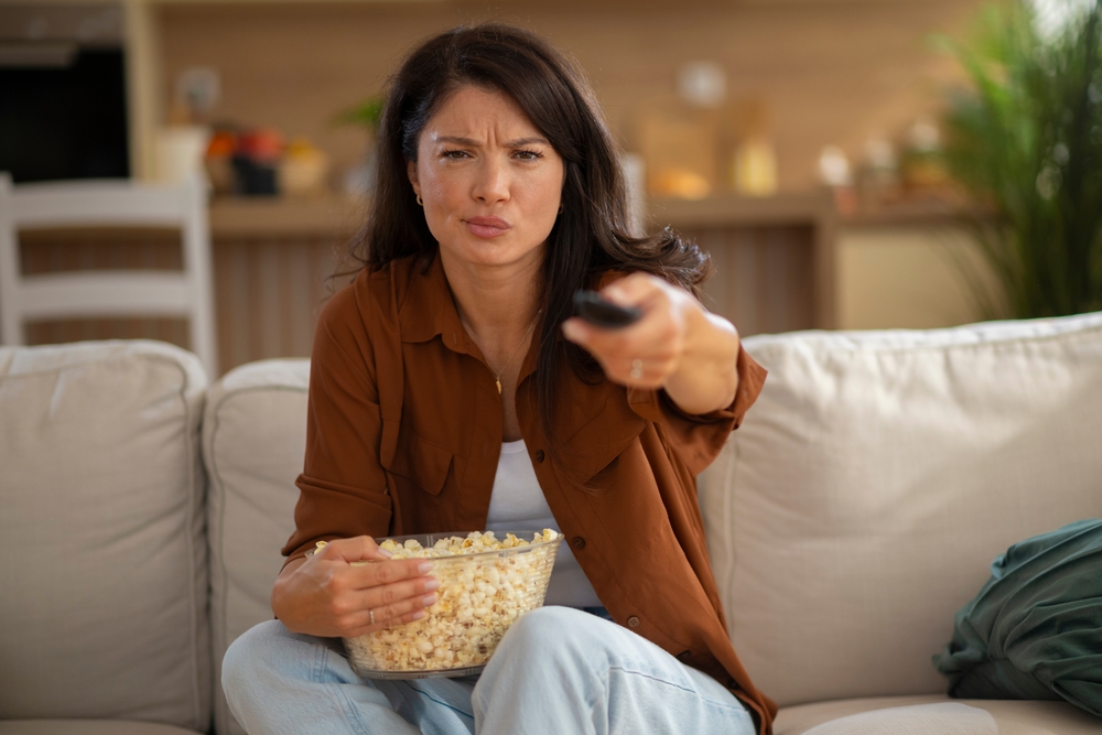 Woman sitting on a couch, watching TV with a focused and slightly annoyed expression while holding a bowl of popcorn.
