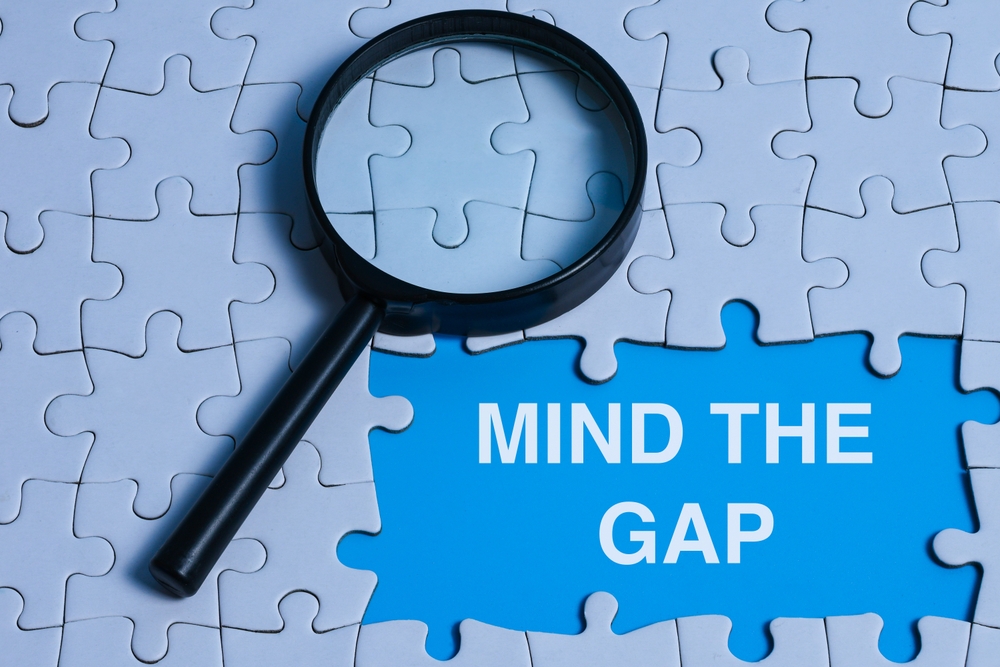 Magnifying glass placed on a puzzle with the text ‘MIND THE GAP’ on the puzzle gap, representing problem solving, critical thinking, and identifying gaps in business or learning