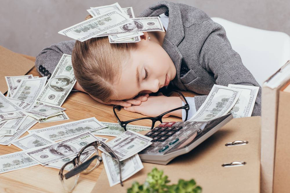a little girl in a suit lying under a pile of money at work isolated on gray