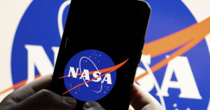 National Aeronautics and Space Administration (NASA) logo, known as the "meatball," which is a blue circle featuring white text, a red vector, and white orbital lines displayed on a smartphone being held by someone