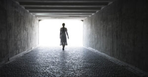 Woman Who Briefly Died Claims to Have Seen the Future—and Issues a Stark Warning silhouette of a person walking towards a bright light at the end of a dark tunnel or corridor