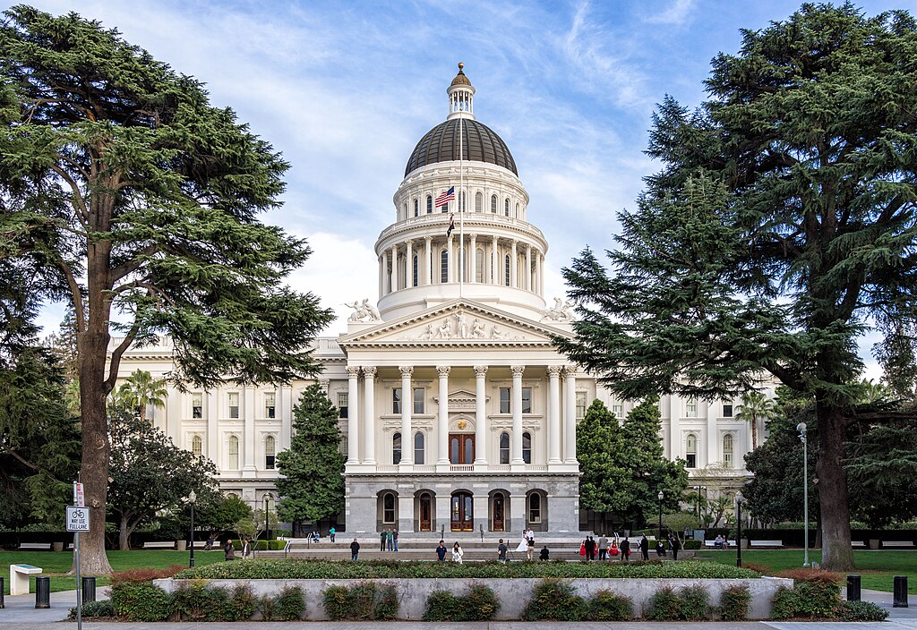 California State Capitol building with its white neoclassical columns and golden dome, framed by tall trees under a partly cloudy sky.