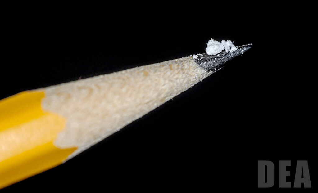 A small cluster of white powder balanced on the sharpened tip of a yellow pencil against a solid black background. This is approximately two milligrams of fentanyl.