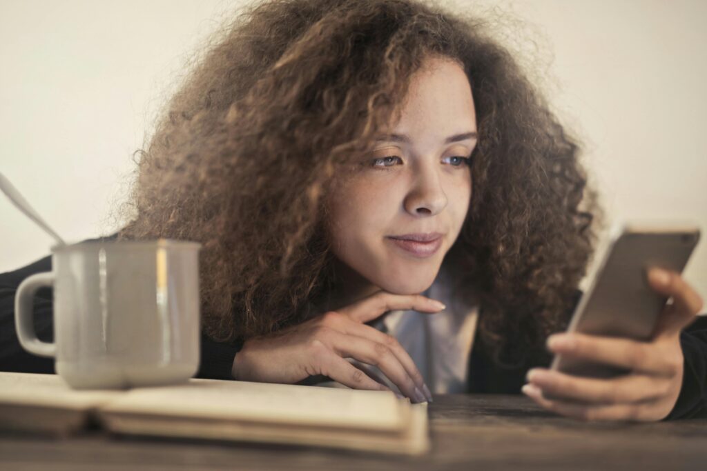 A young woman with curly hair rests her chin on her hand while scrolling through her phone at a table, a coffee mug and open notebook beside her.