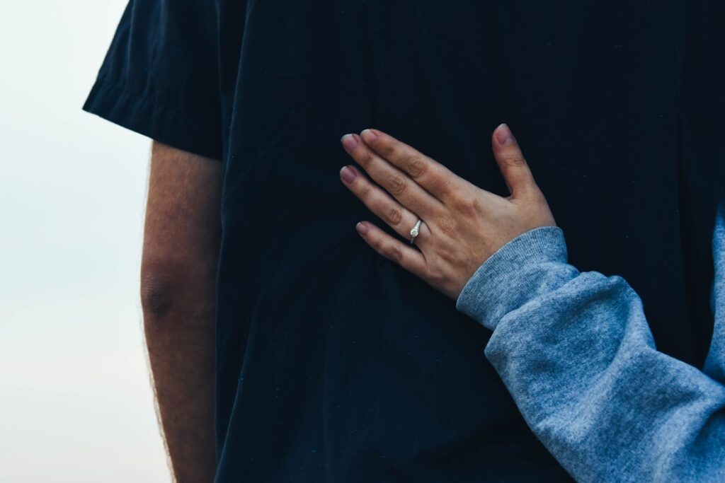 A hand wearing a ring rests gently on someone's back in a gesture of comfort.
