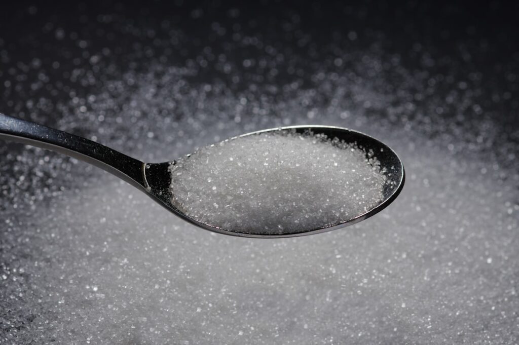 Metal spoon heaped with white granulated sugar, hovering over a surface covered with scattered sugar crystals against a dark background.