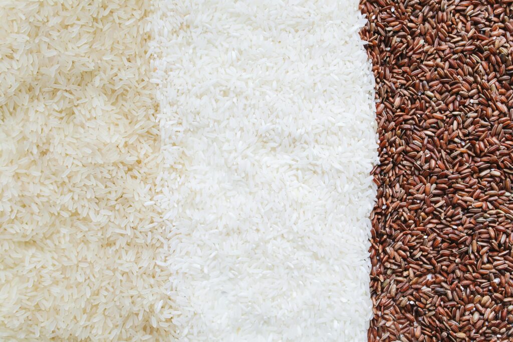Three types of rice displayed side by side, with long-grain white rice on the left, medium-grain white rice in the center, and dark brown or red rice on the right.