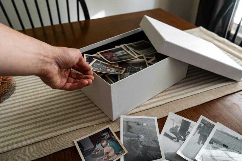 A hand reaches into a white box filled with old photographs, with more black-and-white prints scattered across a table.