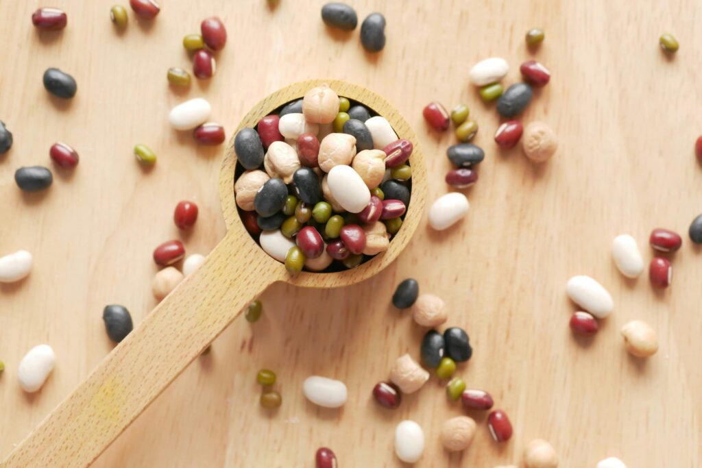 Small wooden spoon overflowing with assorted dried beans and legumes in white, black, red, and green, with more beans scattered across a light wooden surface.