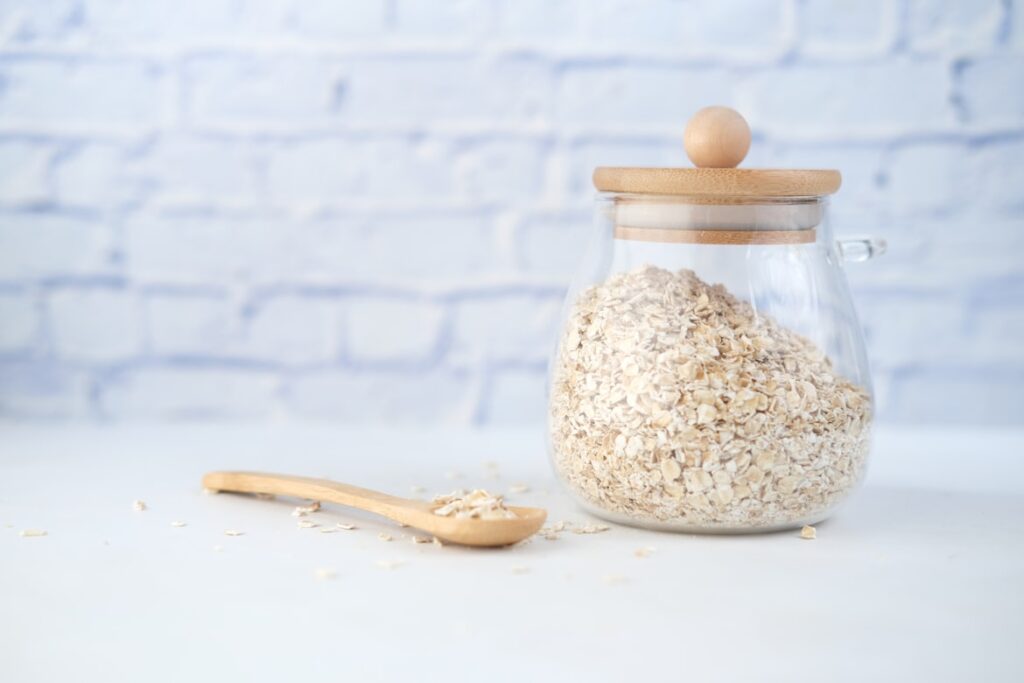 Glass storage jar with wooden lid filled with rolled oats, alongside a wooden spoon, set against a white brick wall.