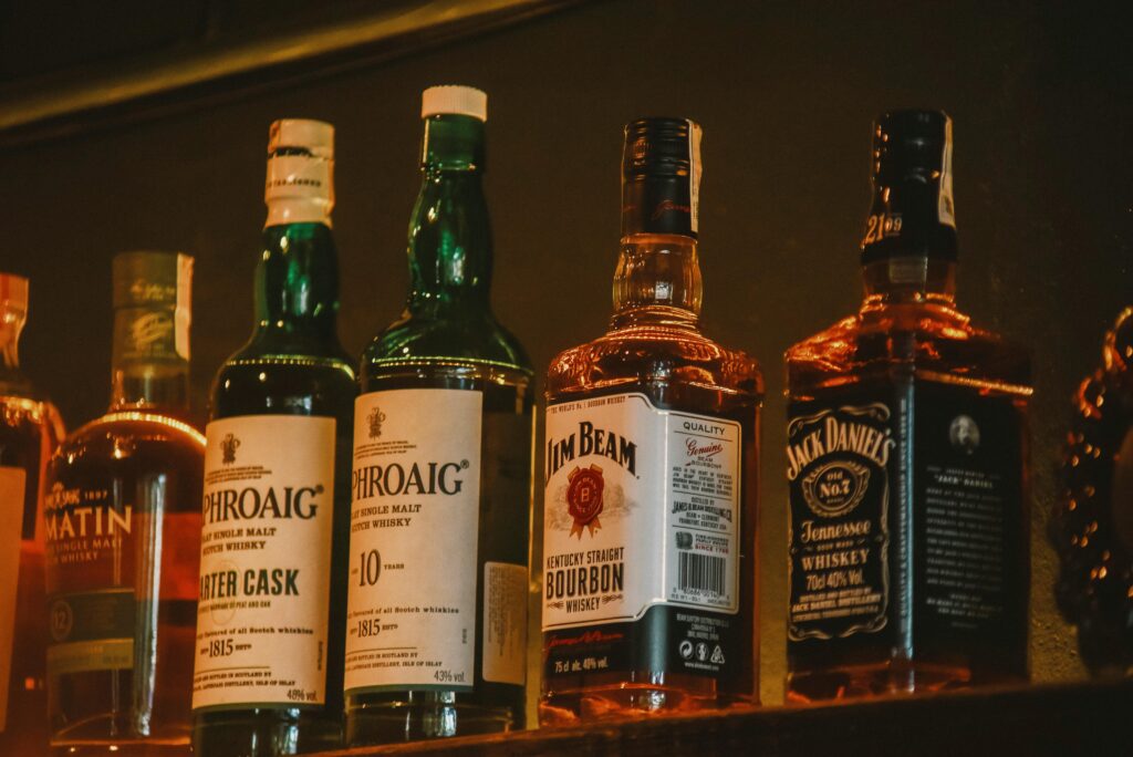 Row of whisky and bourbon bottles on a bar shelf, including Laphroaig, Jim Beam, and Jack Daniel's, warmly lit from behind.
