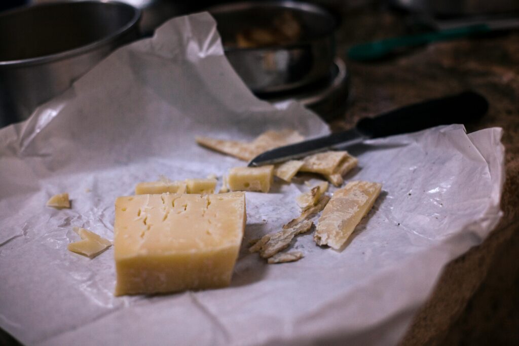 Block of aged hard cheese with broken chunks and crumbs on white parchment paper, with a knife resting nearby on a kitchen counter.