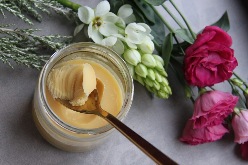 Open glass jar of golden ghee with a spoon scooping some out, photographed from above alongside pink carnations and white flowers on a gray surface.