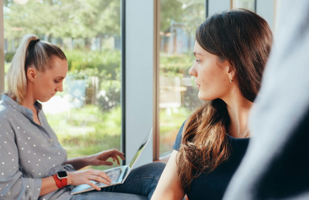 A woman with long brown hair looks back at another woman working on a laptop behind her, her expression quiet and thoughtful as they sit near a window.