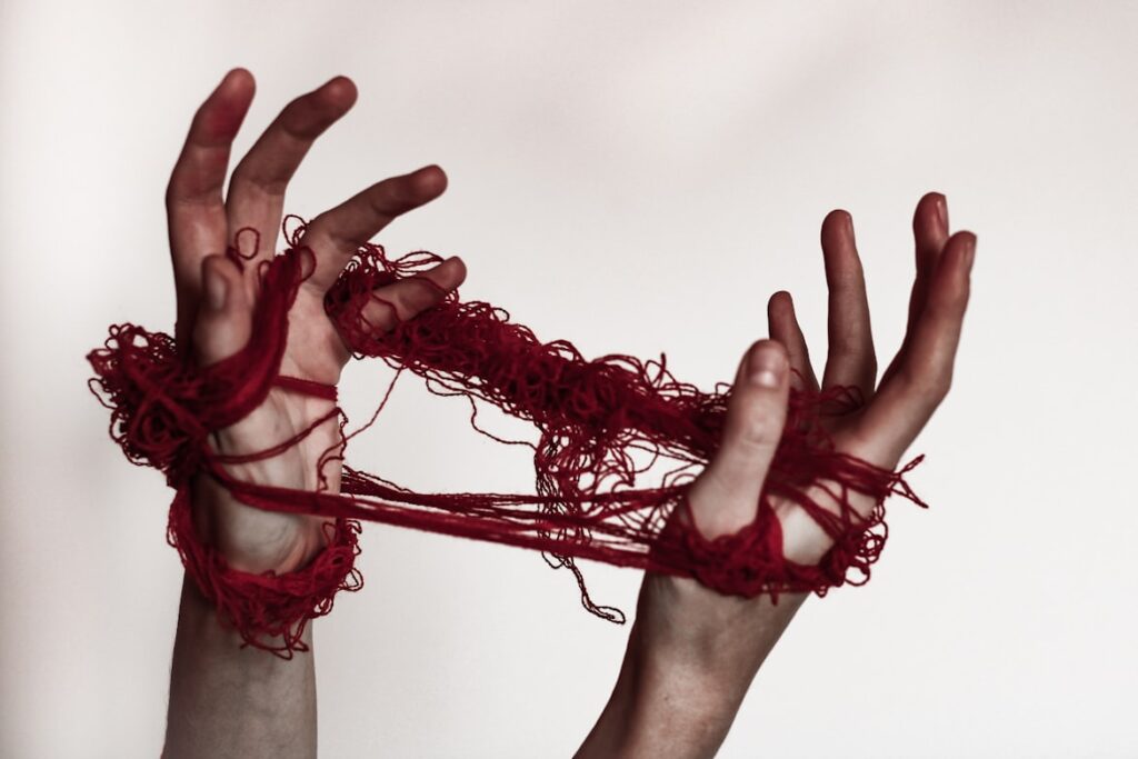 Two hands stretch upward tangled in messy red thread, fingers spread apart as if trying to pull free.