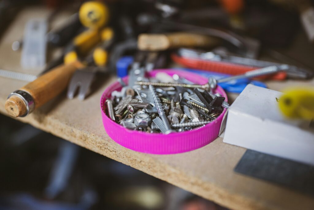 A pink bottle cap filled with assorted screws, nails, and wall anchors sits on a wooden workbench surrounded by tools including a chisel, hammer, and pliers.