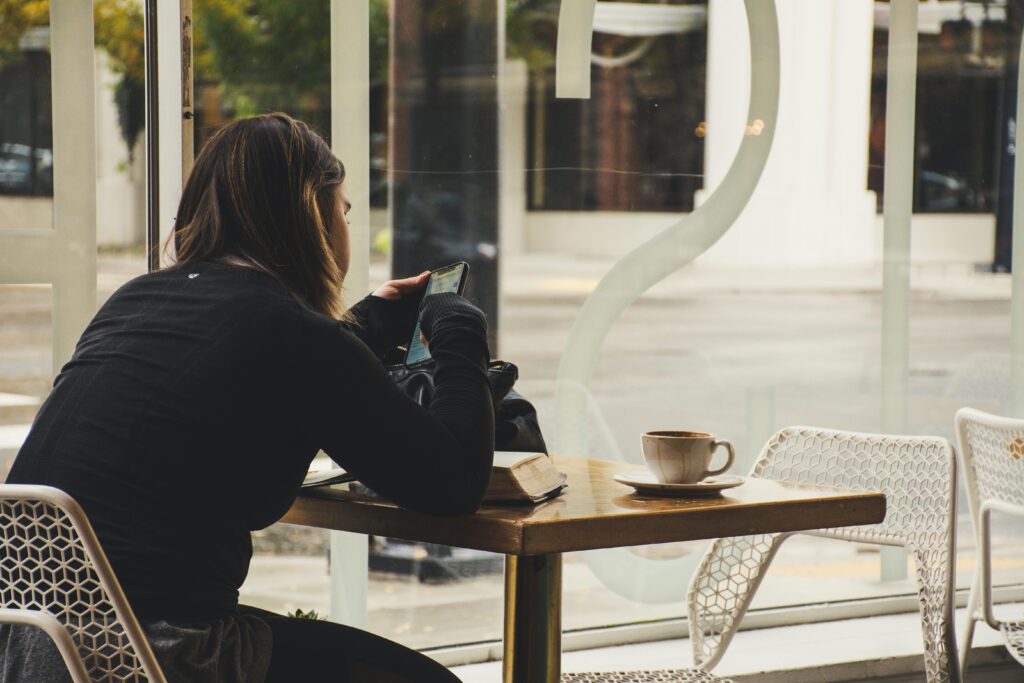 A woman sits alone at a cafe table near a window, holding her phone and gazing outside with a cup of coffee beside her.