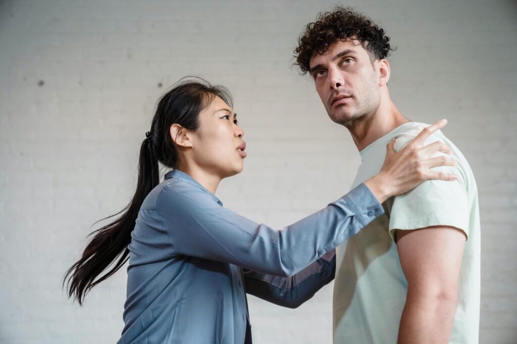 A woman places her hands on a man's shoulders while speaking to him with a serious expression, while the man looks away.