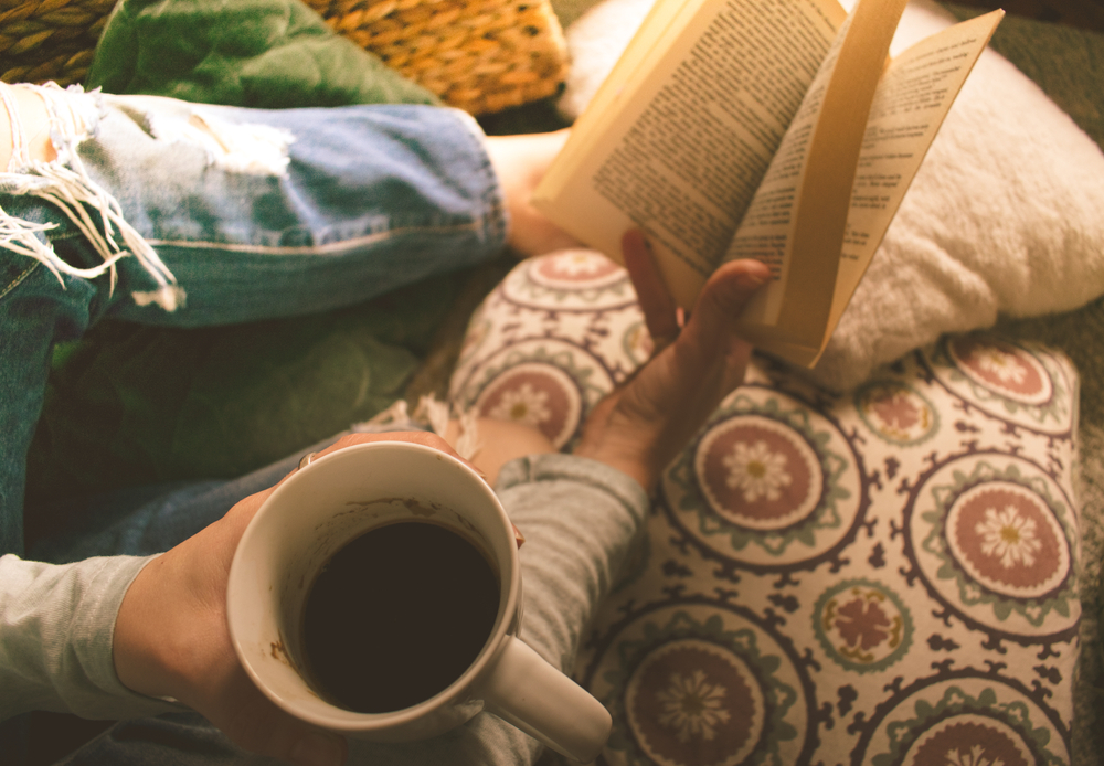 Woman sitting on the floor on decorative pillows in the morning light. Holds a cup of coffee in one hand and a book in the other