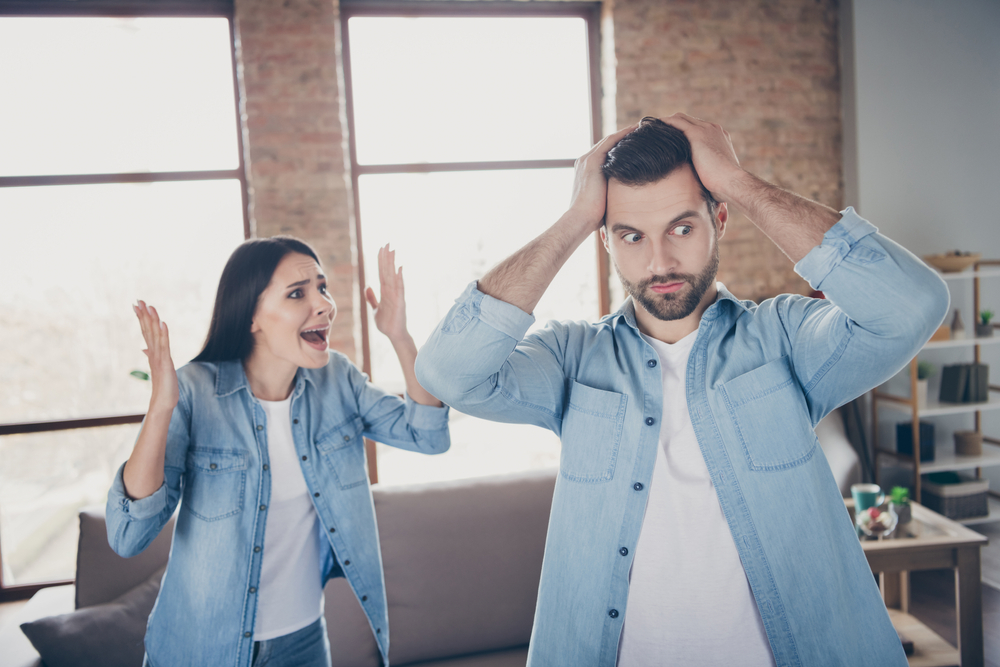 Photo of outraged woman shout man have conflict misunderstanding husband touch hands head astonished wife reaction stand near comfort cozy couch in house indoors