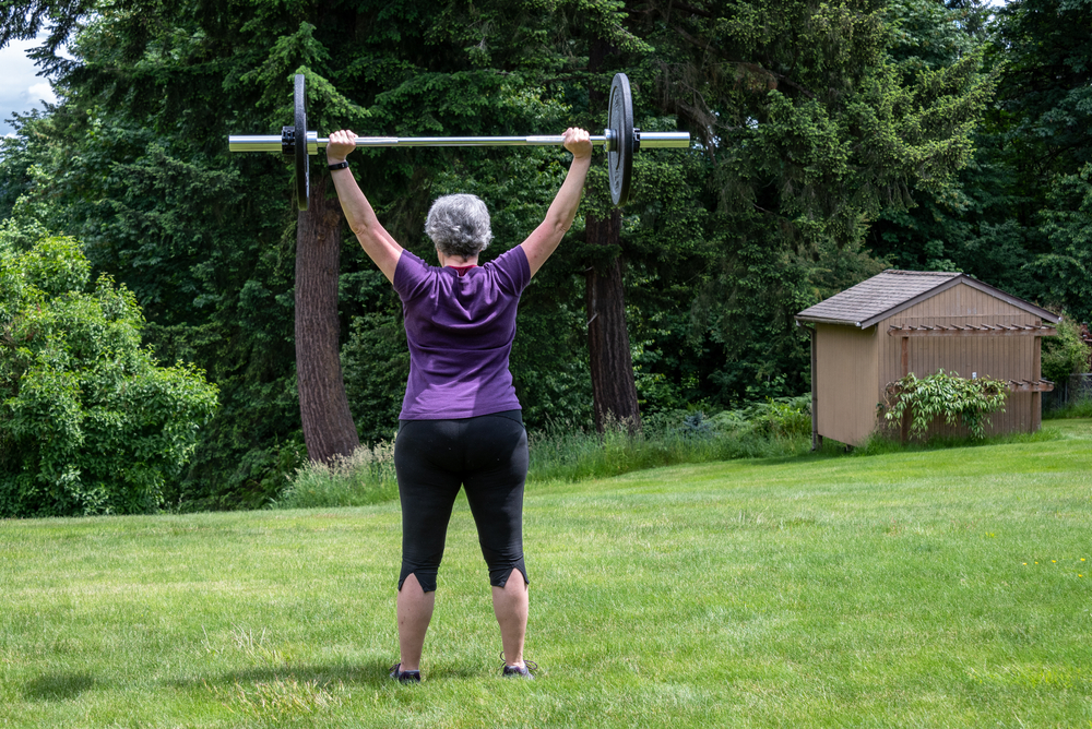 Middle aged caucasian woman with gray hair lifting a barbell with black plates, fitness outside on the lawn
