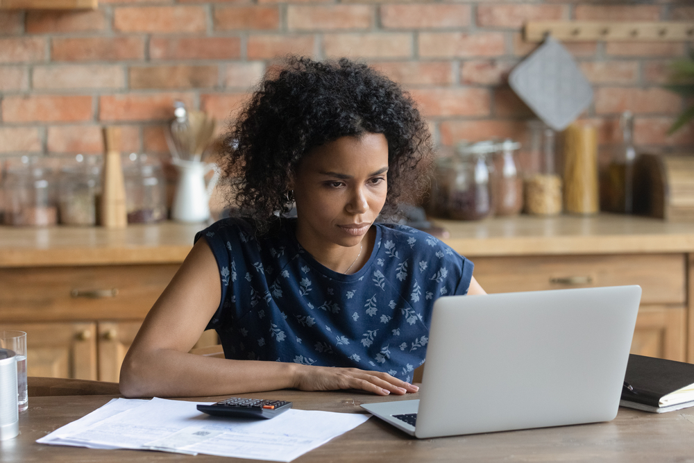 Focused young African American woman sit at desk at home look at laptop screen manage household budget. Serious biracial female calculate finances expenditures, pay bills taxes online on computer.
