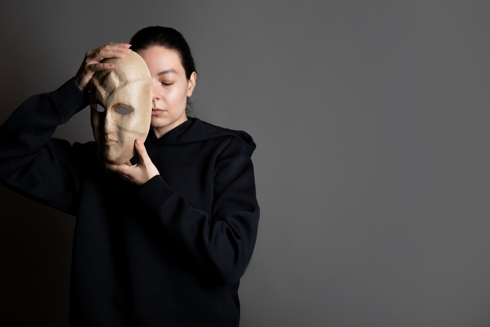 Hiding behind a mask, a young woman in a dark hoodie hides her face with a mask, self-identification problems and impostor syndrome. Portrait in the studio on a dark gray background.