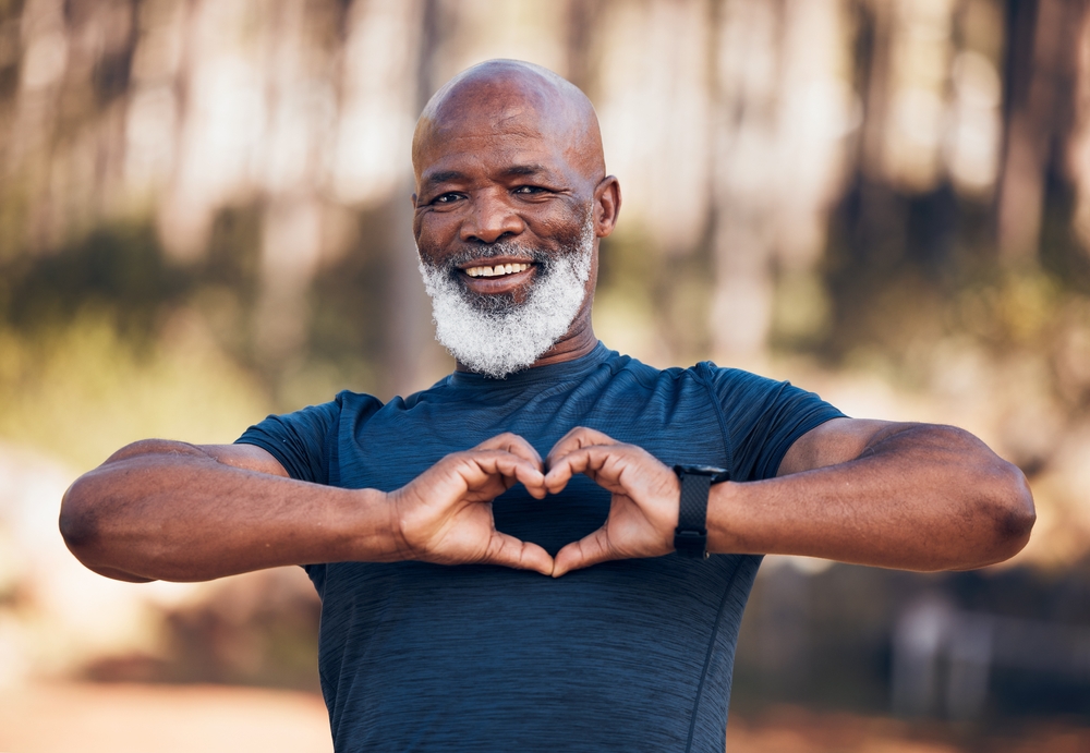 Black man, fitness and heart portrait for exercise, fitness and health while outdoor with a smile. Healthy senior person in nature forest happy with hand emoji sign for workout and cardio training