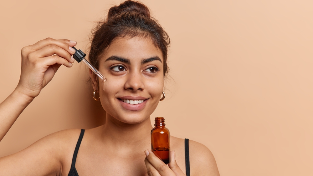 Domestic skin care and anti wrinkle routine. Pleased dark haired woman applies facial serum with dropper smiles toohthily focused aside dressed in t shirt isolated over brown background copy space