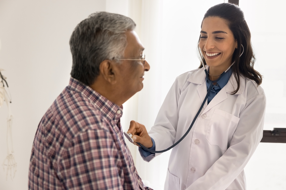 Happy young Latin cardiologist doctor woman listening to heartbeat rate of senior Indian patient, applying stethoscope to chest, speaking, smiling, laughing. Positive cardiologist examining older man