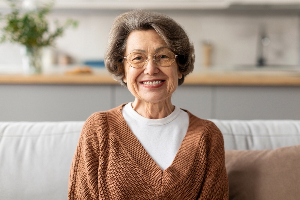 Head shot portrait of happy older European woman wearing eye glasses, looking at camera with toothy smile, posing at home