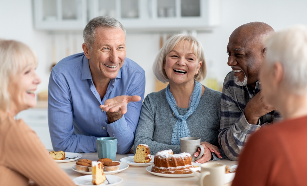 Multiracial group of happy senior people sitting around table drinking tea with cake and having conversation, smiling and laughing, having home party or enjoying time at nursing home