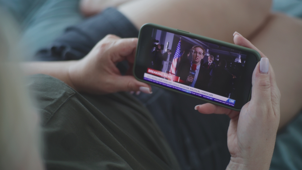 Static shot of adult Caucasian woman watching online breaking news report about the elections in the United States using mobile phone while lying on sofa at home. Election Day in USA. Lifestyle